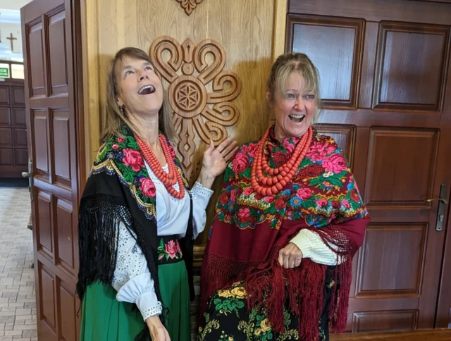 Two women in colorful traditional attire with floral shawls and bead necklaces pose joyfully indoors in front of a wooden wall.