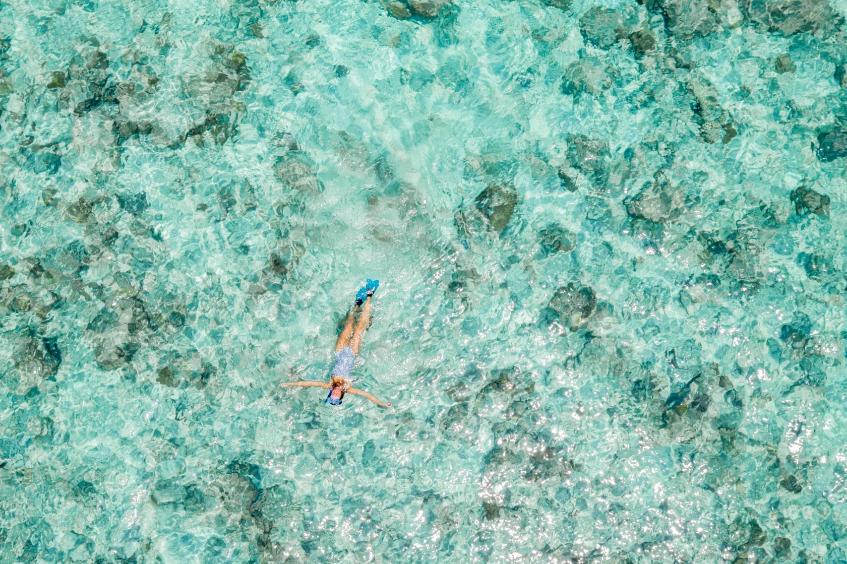 A person snorkeling in clear turquoise water, surrounded by scattered rocks and sand, viewed from above.