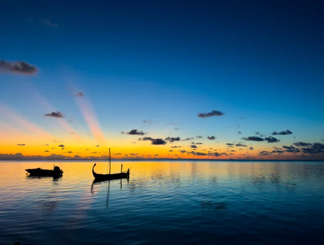 Two boats float on a calm sea at sunset, with a clear sky and a warm glow on the horizon.