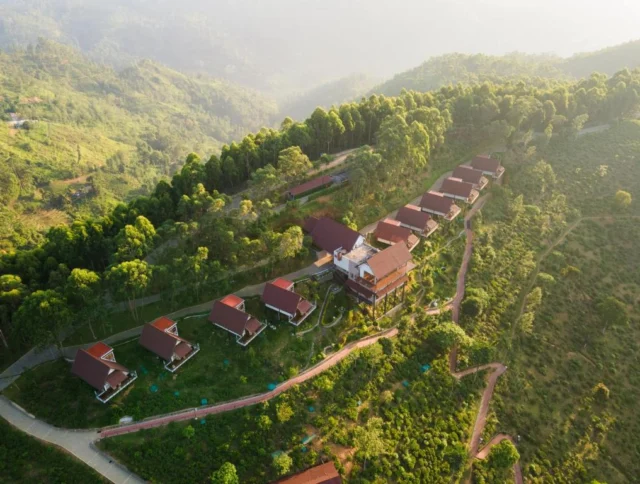 Aerial view of a hillside resort with red-roofed cabins surrounded by lush greenery and trees. Paths connect the cabins on the slope.