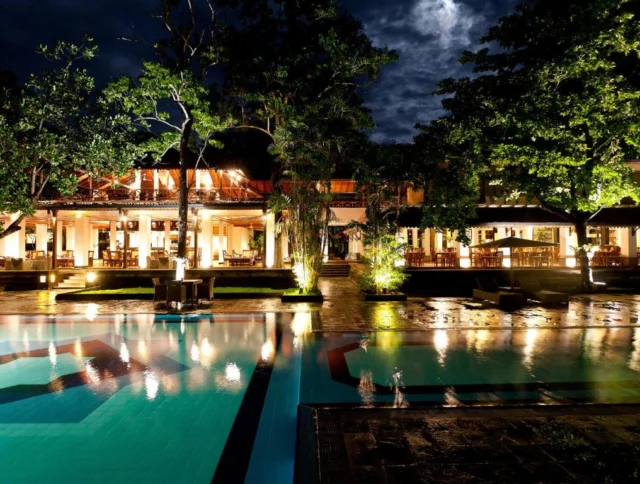 A nighttime view of a lit-up building with a pool in the foreground, surrounded by trees and illuminated by various lights.