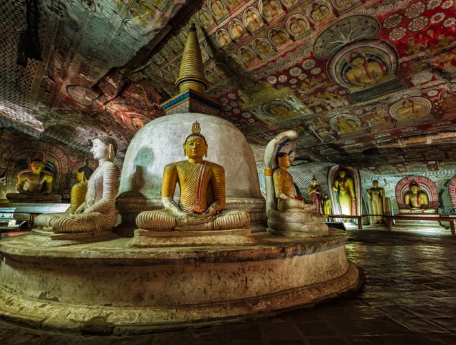 Buddha statues and colorful murals inside an ancient, ornately decorated cave temple.