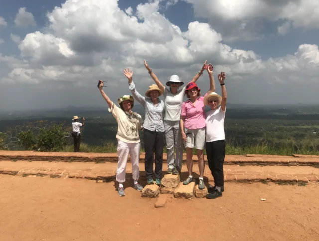 Five people with hats and sunglasses raise their arms joyfully on a dirt path with a scenic view behind them. A person stands in the distance taking a photo.