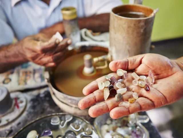 Hand holding various uncut gemstones near a polishing wheel with another person adjusting a stone.