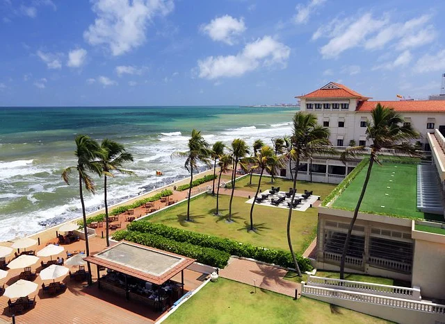 Oceanfront view with palm trees, a large building with a red roof, green lawn, and outdoor seating area overlooking the waves under a bright blue sky.