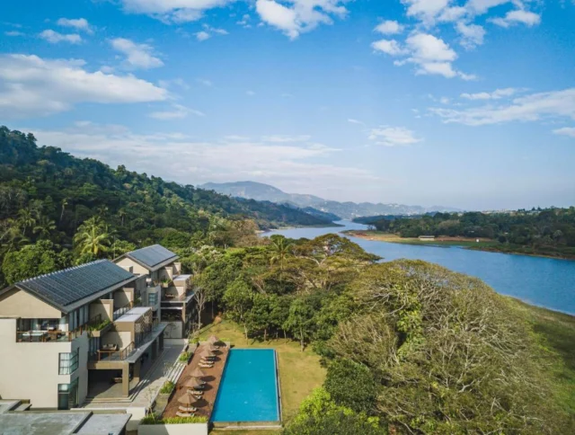 Aerial view of a modern building with a pool, surrounded by lush greenery and a river, set against a backdrop of hills and a partly cloudy sky.