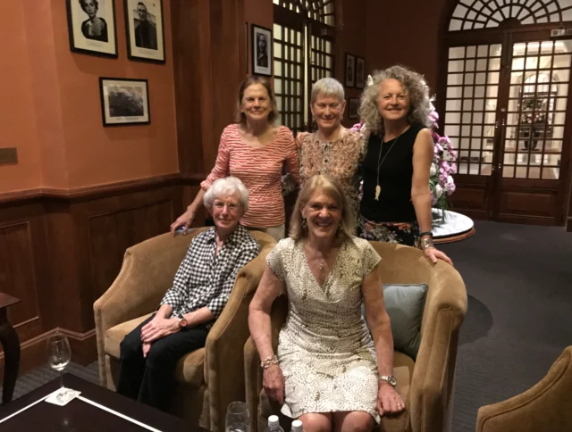 Five women are posing in a warmly lit room. Two are seated on chairs, and three are standing behind them. Framed pictures are on the walls.