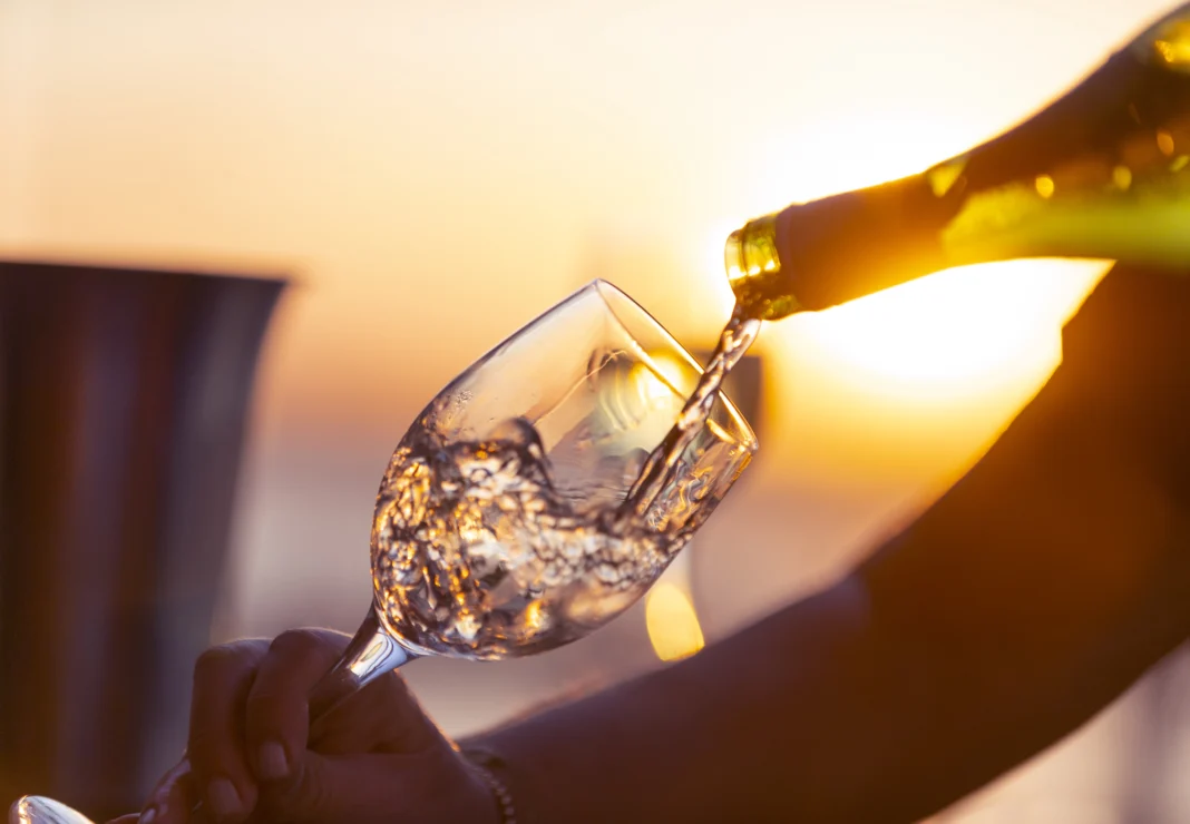 A hand pours white wine from a bottle into a glass against a sunset backdrop.