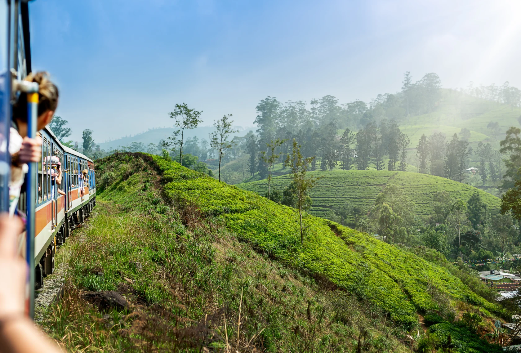 A train travels through lush green hills with passengers leaning out for a view.