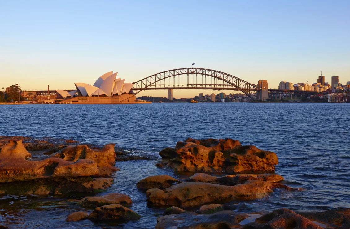 View of Sydney Opera House and Harbour Bridge at sunset, with rocky foreground and city skyline in the background.