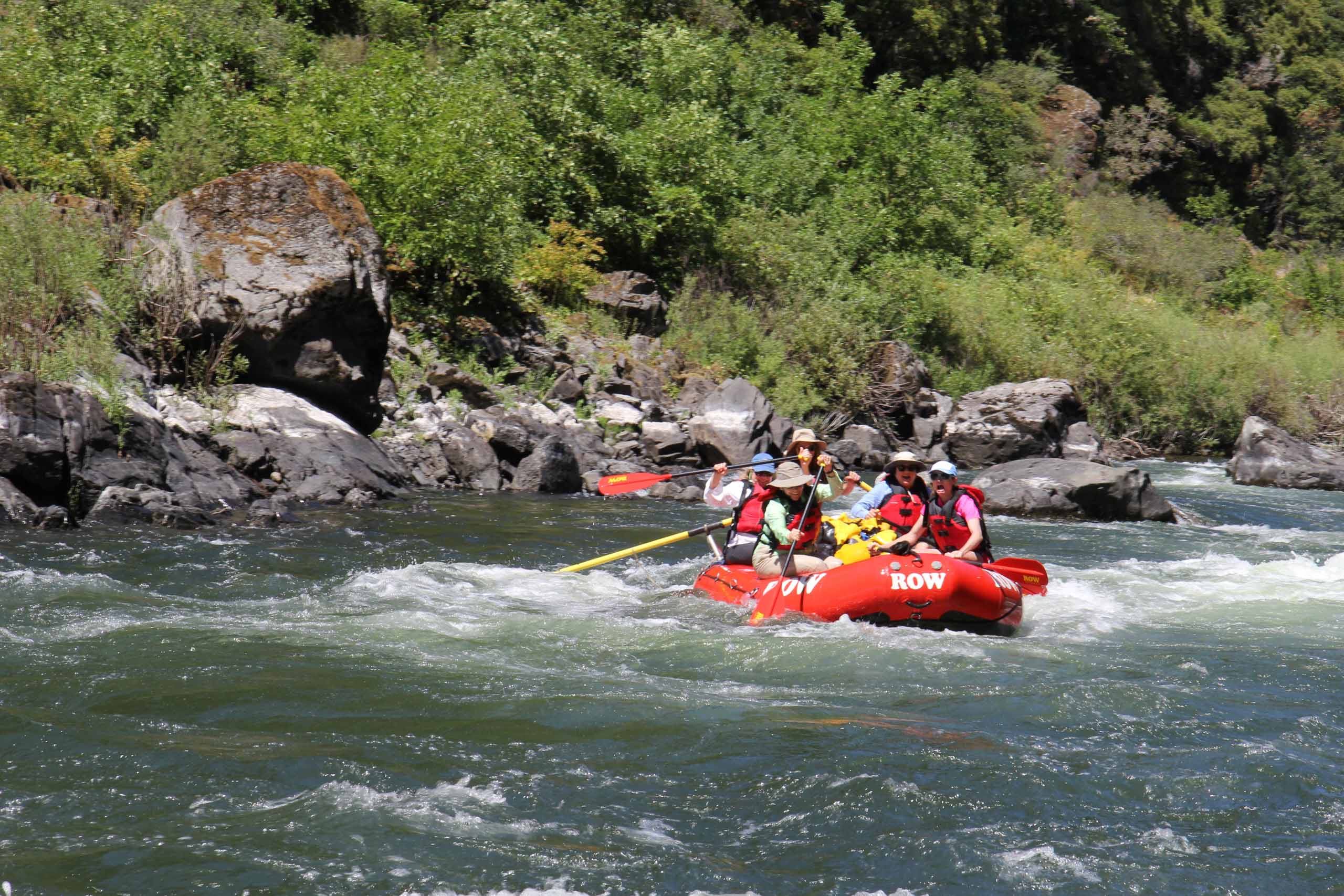 A group of people in helmets and life jackets are white-water rafting in a red inflatable raft on a river surrounded by lush greenery and rocks.