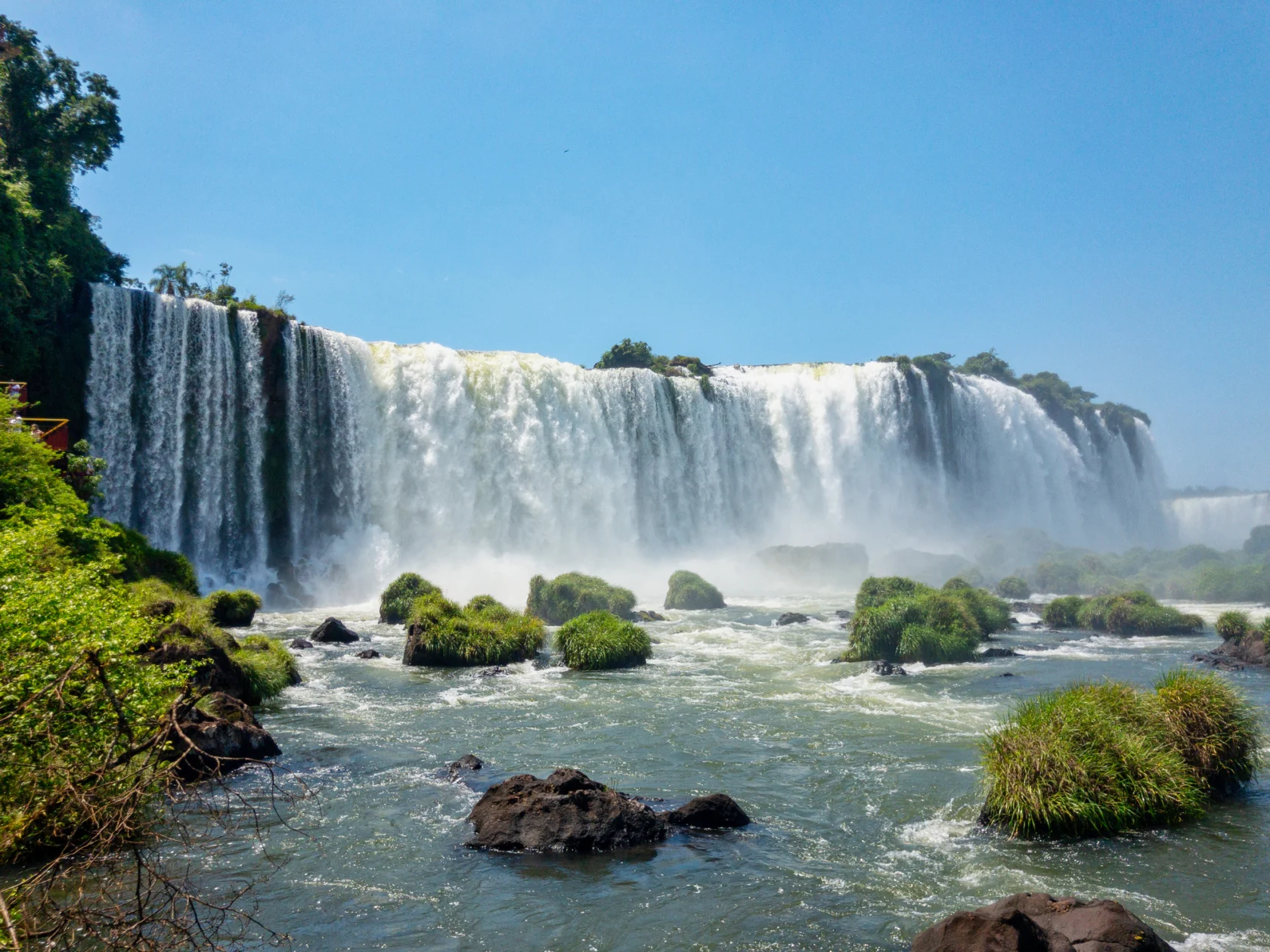 Waterfalls cascading over a cliff into a river, surrounded by lush greenery and rocks under a clear blue sky.