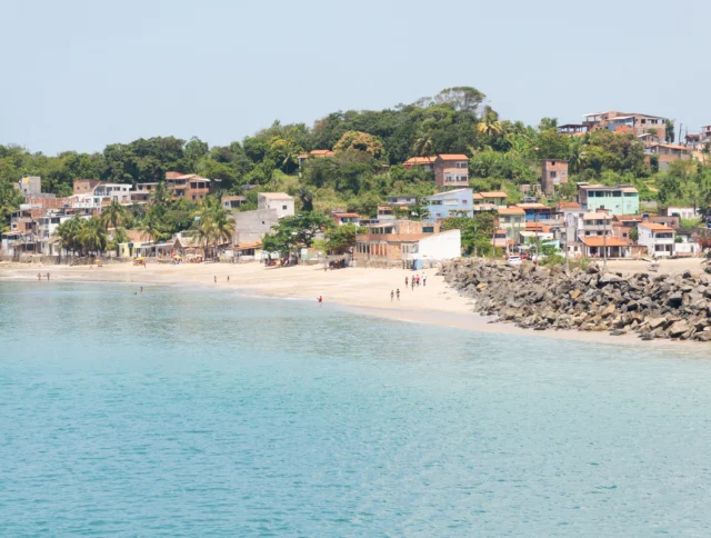 Coastal view of a beach with clear blue water, people walking, and houses surrounded by greenery in the background.