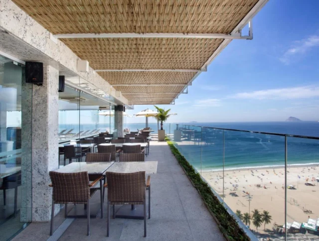 Outdoor terrace with wicker furniture overlooking a sunny beach and ocean. Glass railing and awning provide unobstructed views.