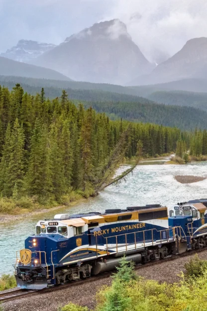 A blue and gold train travels through a forest alongside a river, with misty mountain peaks in the background.