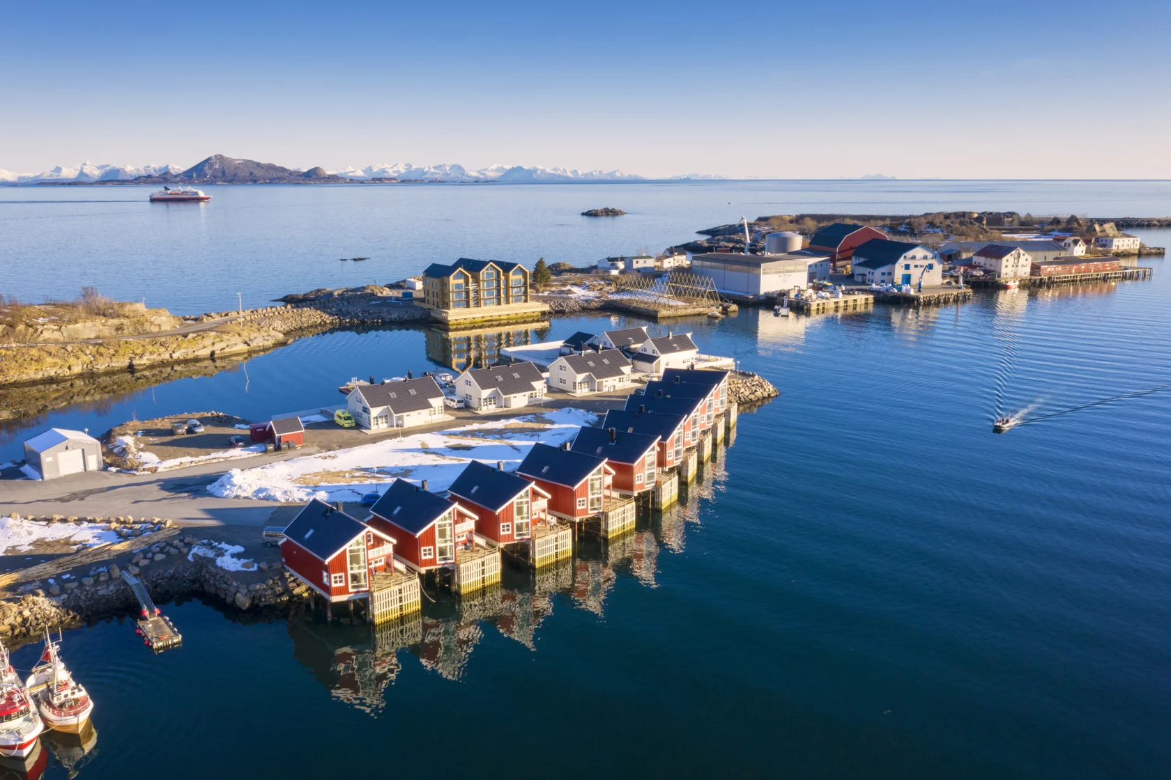 Coastal village with red buildings over water, snowy mountains in the background, boats, and a ship in the distance under a clear sky.