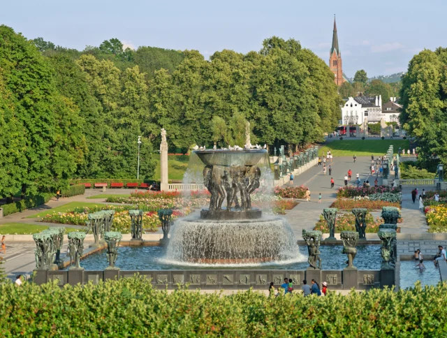 A fountain with sculptures is surrounded by trees and walkways in a park, with a church spire visible in the background. People are walking and sitting around the area.