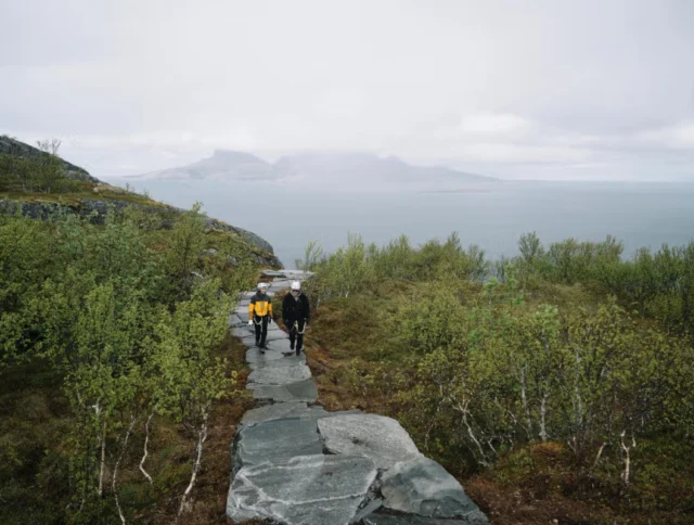 Two people walk on a stone path surrounded by greenery, overlooking a misty body of water and distant mountains.