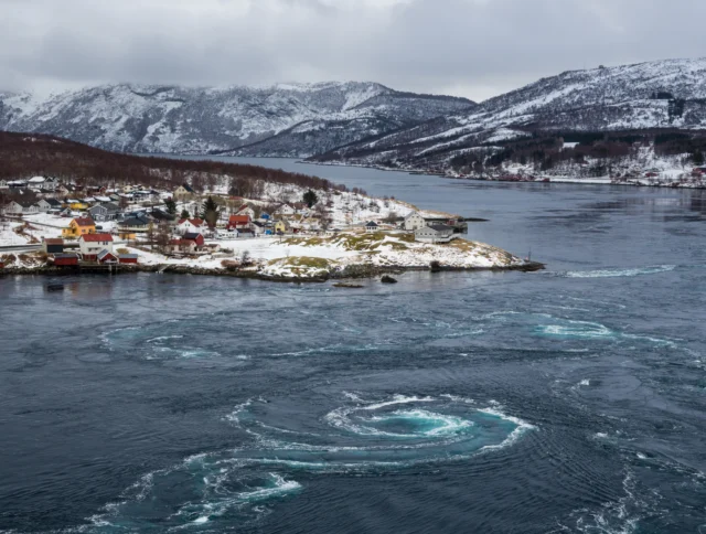 Aerial view of a snowy coastal village with houses near a fjord. Swirling water patterns are visible in the foreground, and mountains are in the background under a cloudy sky.