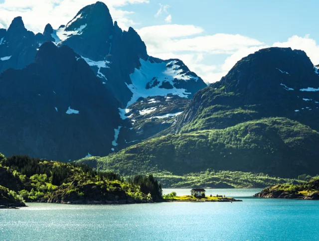 A solitary house is nestled on a small island surrounded by a blue lake, with towering mountains in the background under a partly cloudy sky.