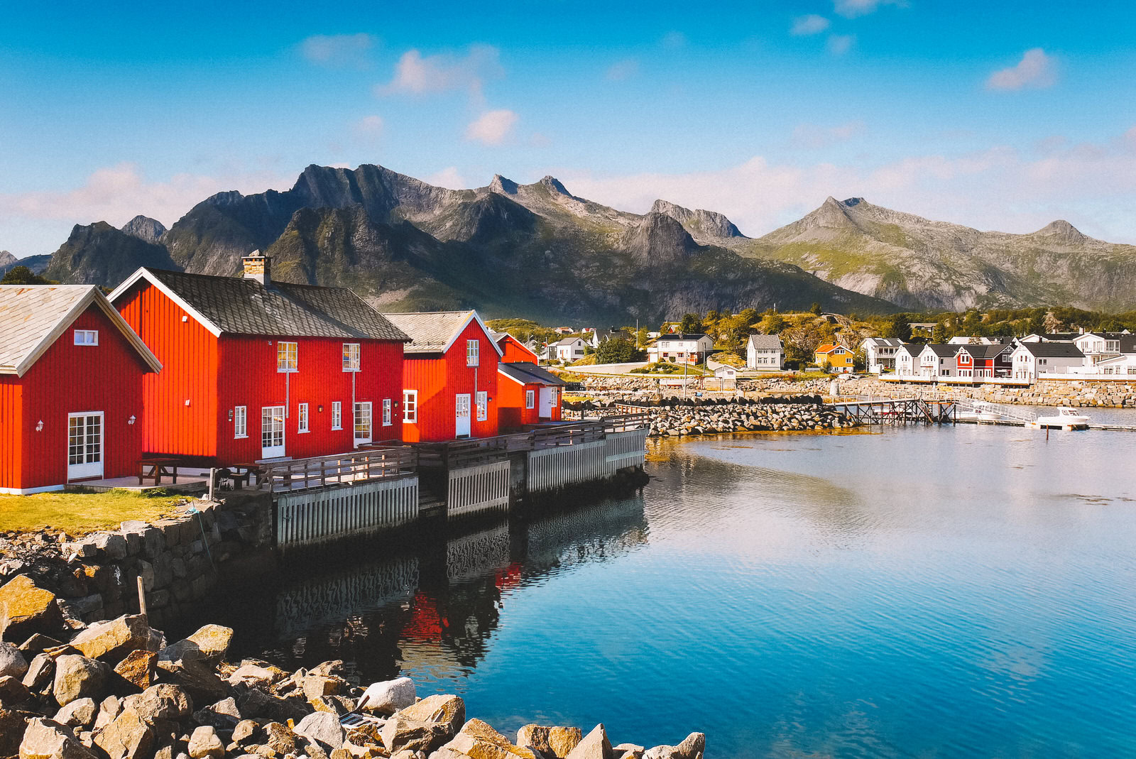 A coastal scene with red wooden houses by a calm body of water, rocky shoreline, and mountains under a clear blue sky.