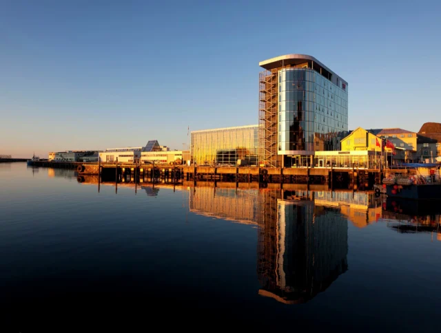 Modern glass building reflecting on calm water at a waterfront in evening light, surrounded by smaller structures and a partially visible mountain in the background.