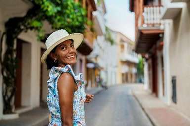 Woman wearing a hat and floral dress smiles while walking on a narrow, plant-lined street.