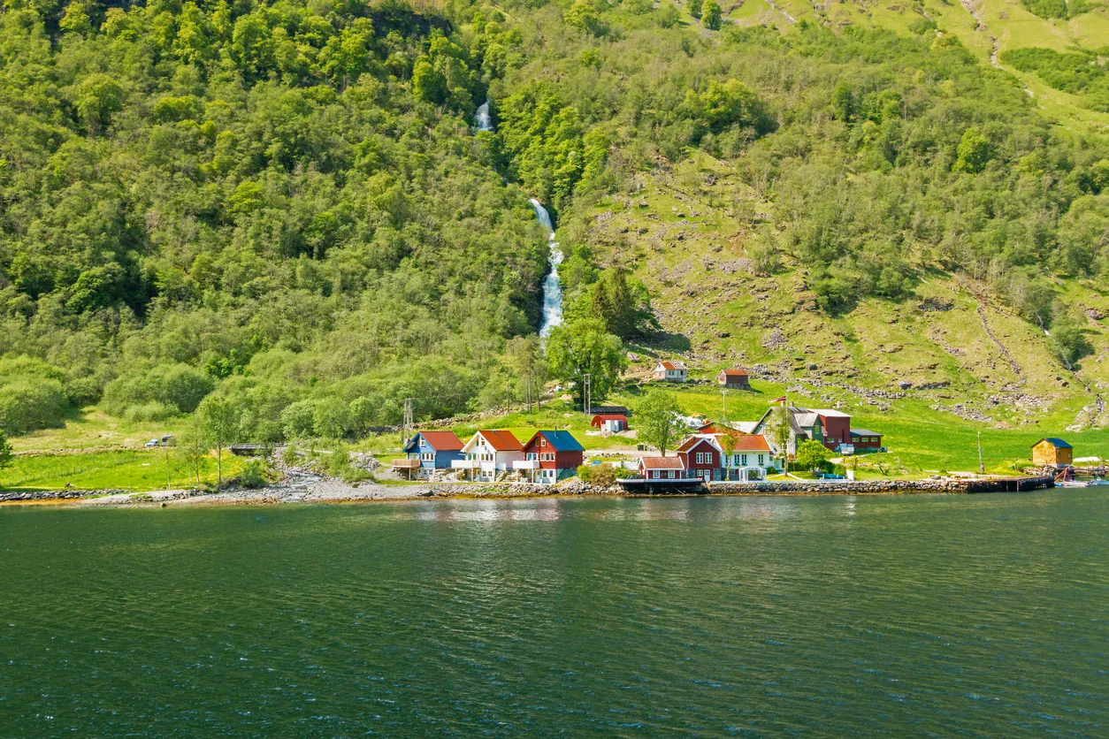 Small cluster of colorful houses by a calm fjord, with a waterfall and green, tree-covered hillside in the background.