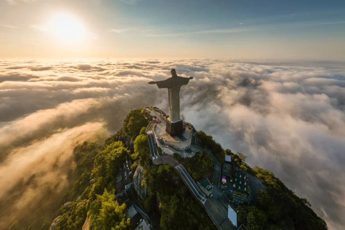 Aerial view of a large statue on a mountain surrounded by clouds with a setting sun in the background.