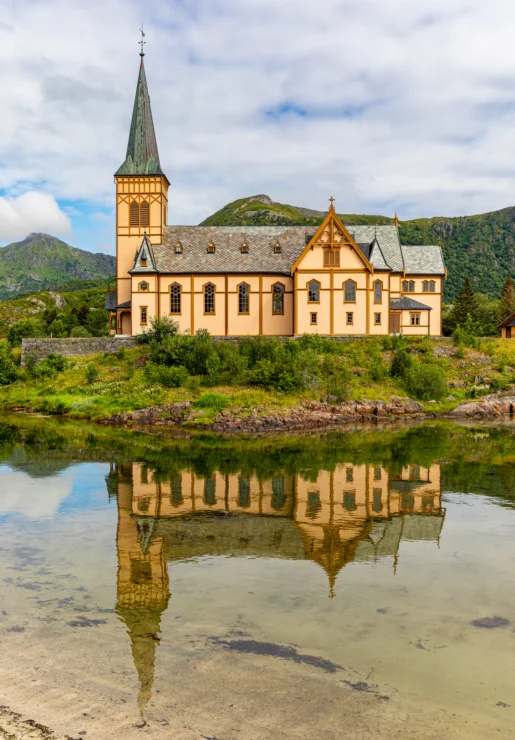 A yellow church with a tall steeple is reflected in a calm body of water, set against a backdrop of green hills under a partly cloudy sky.