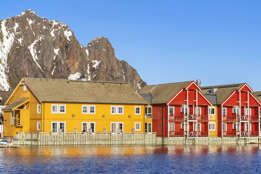 Colorful houses with yellow and red facades are reflected in calm water, set against a backdrop of rocky, snow-dusted mountains under a clear blue sky.