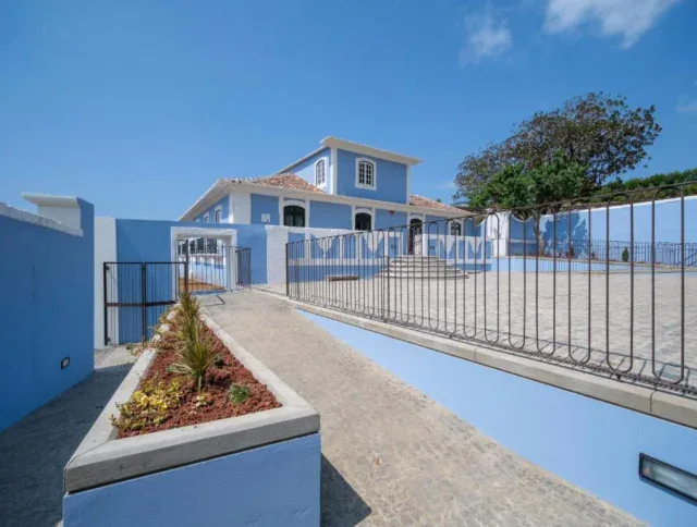 A light blue two-story house with white trim, surrounded by a paved courtyard, a ramp with plants, metal railings, and a tree in the background under a clear sky.