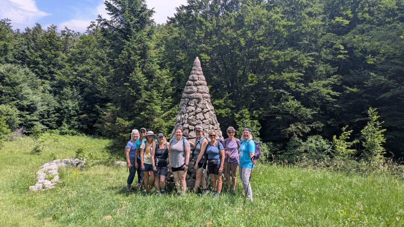 A group of people stand in front of a stone cairn in a grassy clearing, surrounded by trees on a sunny day.