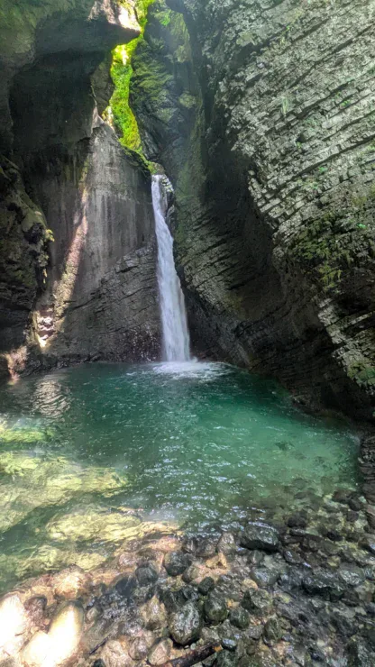 A waterfall flows into a clear turquoise pool surrounded by rocky cliffs and greenery, with sunlight filtering through above.