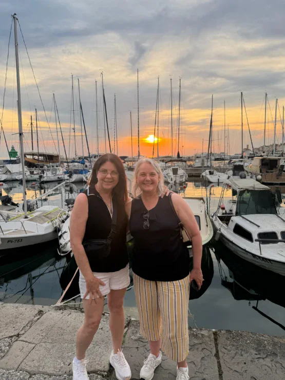 Two women standing and smiling at a marina with sailboats, with the sun setting in the background and calm water reflecting the sky.