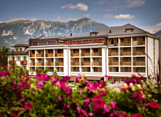 A multi-story hotel with balconies stands in front of a mountain range, with pink and purple flowers in the foreground under a partly cloudy sky.
