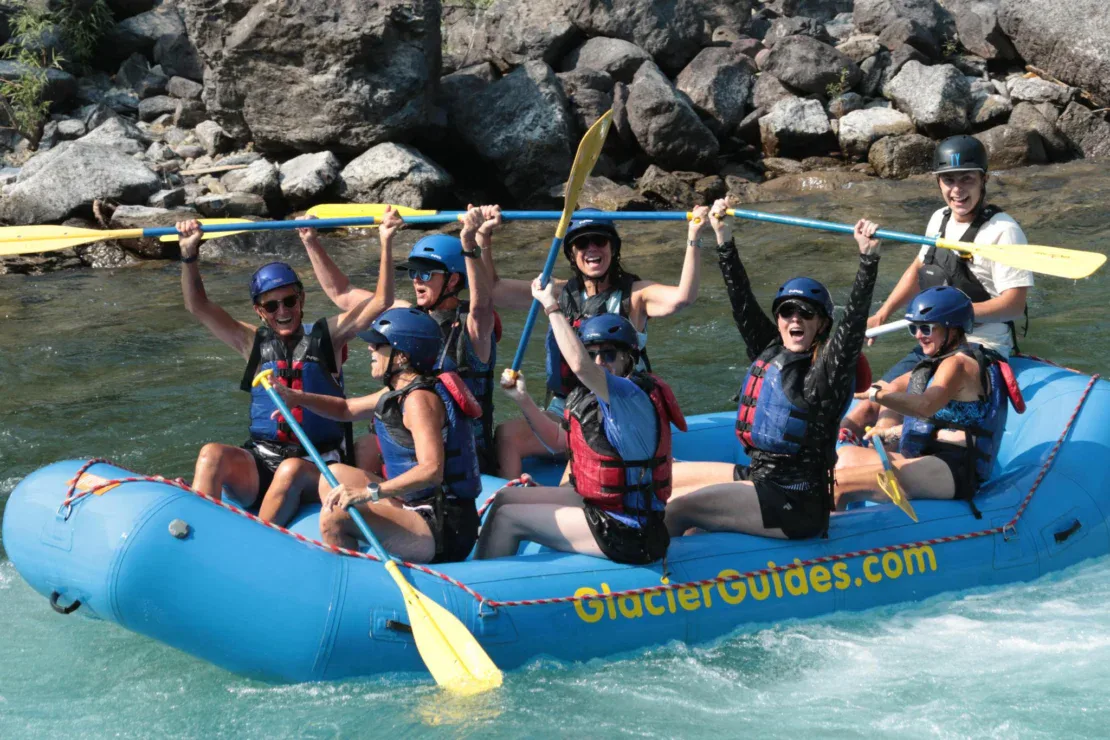 A group of eight people in helmets and life jackets are whitewater rafting on a river in a blue raft labeled “GlacierGuides.” Several raise paddles and smile. Rocks are visible behind them.