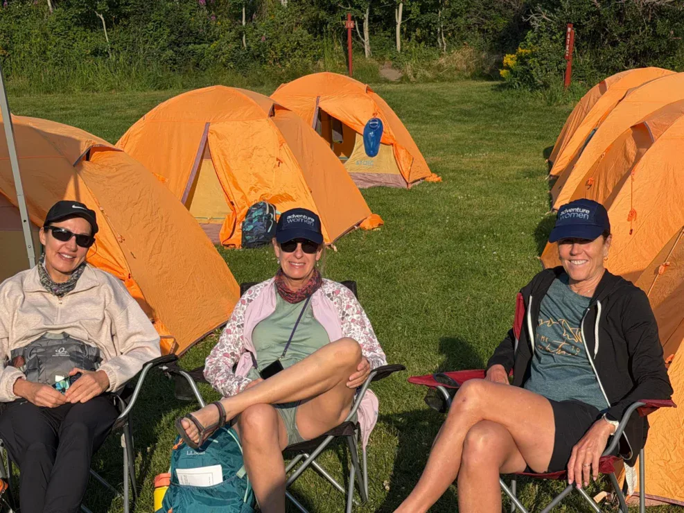 Three women sit in camp chairs on grass in front of several orange tents, wearing hats and sunglasses, enjoying a sunny day outdoors.