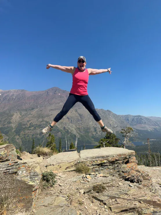 A person in athletic clothes jumps with arms and legs outstretched on a rocky ledge, with mountains and a clear blue sky in the background.