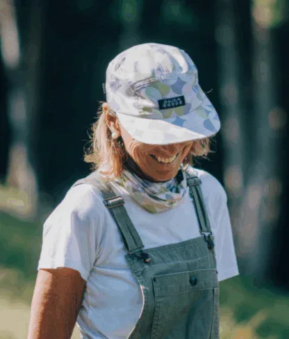 A person wearing a patterned cap, white t-shirt, and green overalls stands outdoors, smiling with their head slightly lowered.