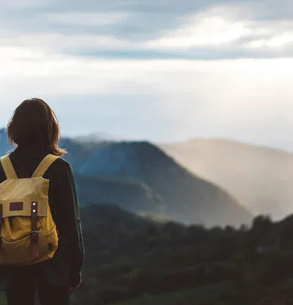 Person with a yellow backpack stands facing a mountainous landscape under a cloudy sky with sunlight breaking through.