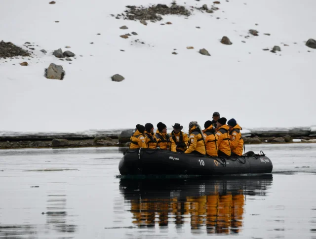 A group of people wearing yellow jackets sit in a black inflatable boat on calm water near a snowy, rocky shoreline.