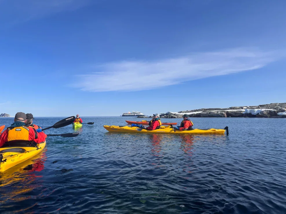 Several people in yellow kayaks paddle on calm blue water near rocky, snow-capped shores with a large ship visible in the background under a clear sky.