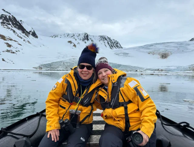 Two people in yellow jackets sit on a boat in front of snowy mountains and a glacier, wearing winter hats and holding cameras.