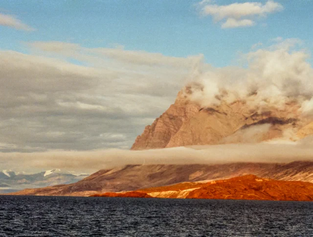 A large, rocky mountain partially covered by clouds rises behind a misty shoreline, with calm blue water in the foreground.