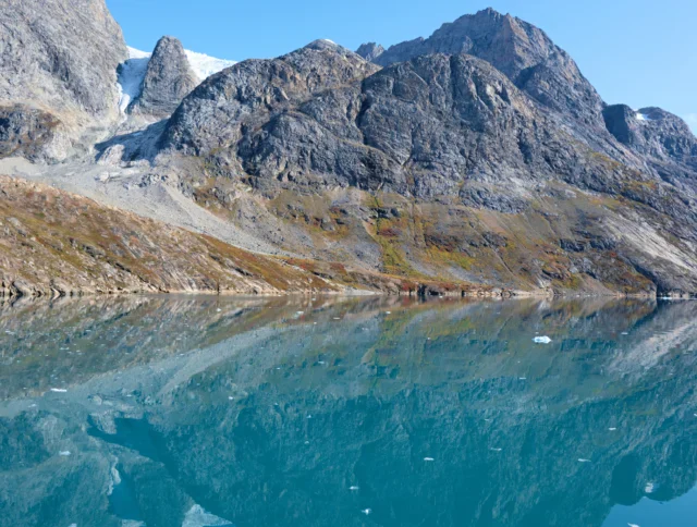 Clear blue lake with floating ice pieces reflects rocky mountains and partially snow-covered peaks under a clear sky.