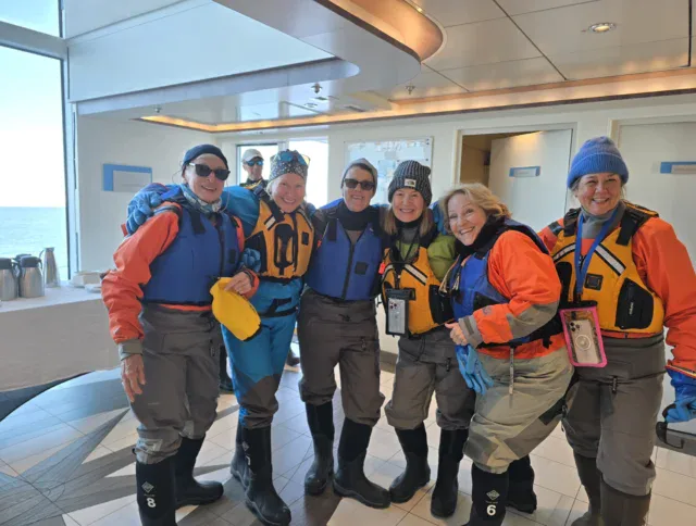 Six people wearing winter hats, life jackets, and waterproof gear stand together indoors on a cruise or research vessel, posing and smiling for the camera.