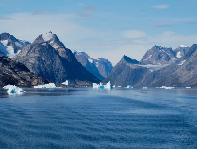 A calm body of water with several icebergs floating, surrounded by rugged, snow-capped mountains under a partly cloudy sky.