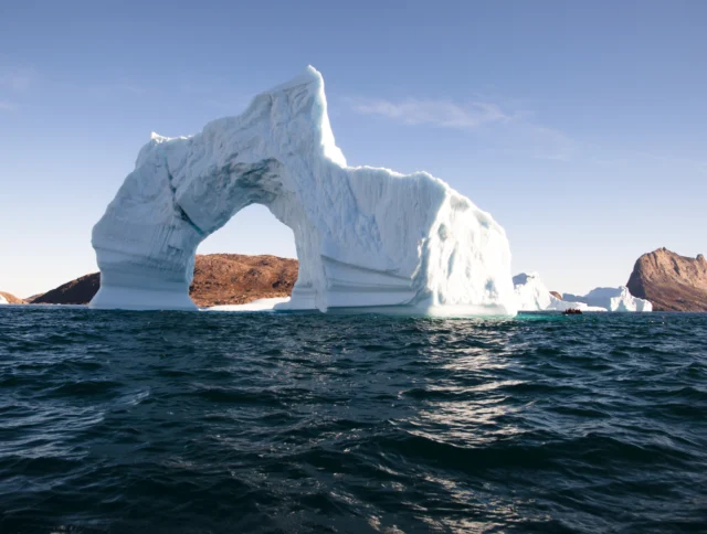 Large iceberg with a natural arch floats in calm blue water, with rocky land and clear sky in the background.