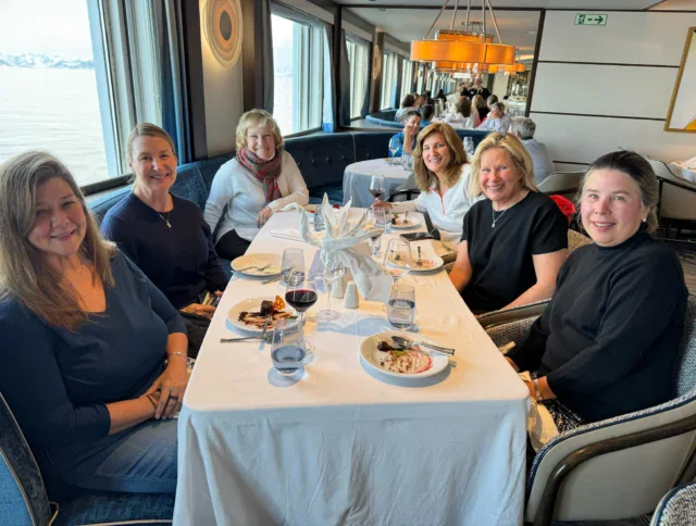 Six women sit around a rectangular table with plates of dessert and drinks in a well-lit restaurant with large windows.
